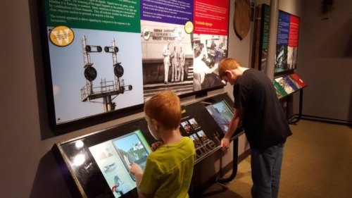 Two children interact with touchscreen displays in a museum, surrounded by informational panels about trains.