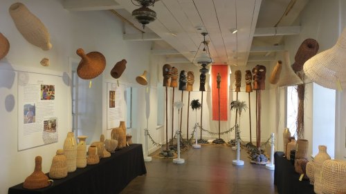 A gallery showcasing woven artifacts, including masks and baskets, displayed on tables and walls in a bright, open space.