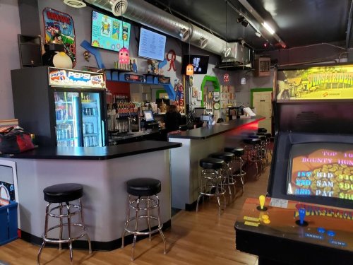 A retro arcade bar featuring a long counter, arcade machines, and colorful decor with vintage gaming memorabilia.