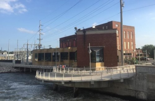 A brick building with large windows sits by a river, featuring a curved walkway and power lines in the background.