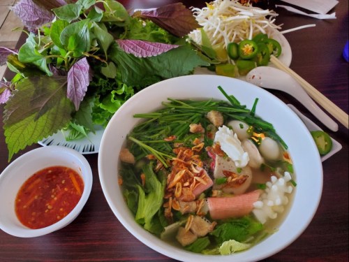 A bowl of soup with noodles, herbs, and meat, accompanied by fresh vegetables and a small dish of dipping sauce.