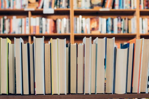 A row of closed books on a table, with a blurred bookshelf filled with books in the background.