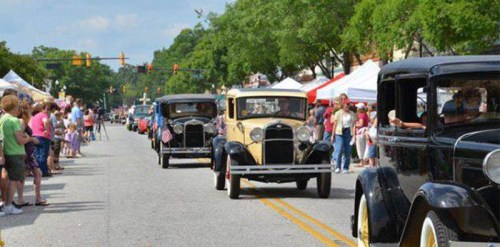A parade of vintage cars drives down a street lined with spectators and colorful tents under a blue sky.