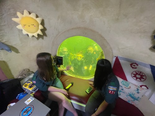 Two people interact with a glowing underwater scene through a circular window, surrounded by colorful decor.
