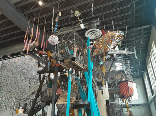 A colorful art installation featuring hanging sculptures, birdcages, and a large fish, set in a spacious gallery.