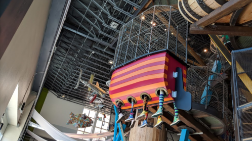 Colorful play structure resembling a ship, with striped sides and children's legs hanging down, set in a vibrant indoor space.