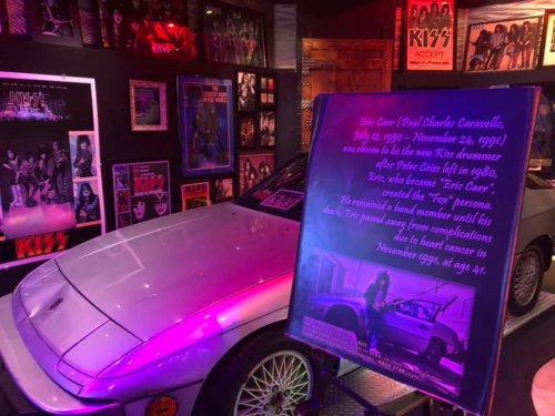 A display featuring a car and a tribute sign for Eric Carr, former drummer of KISS, with band posters in the background.