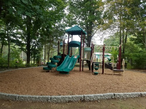 A playground with green slides and climbing structures, surrounded by trees and wood chip ground cover.