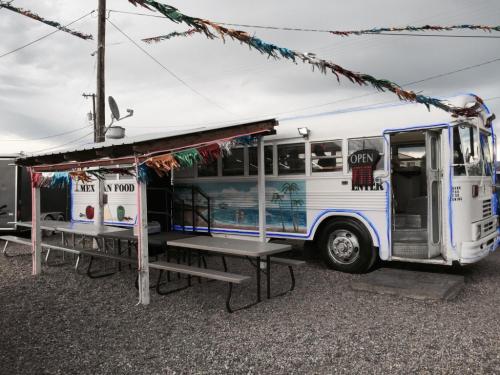 A colorful food truck styled as a bus, with outdoor seating and festive decorations, set against a cloudy sky.