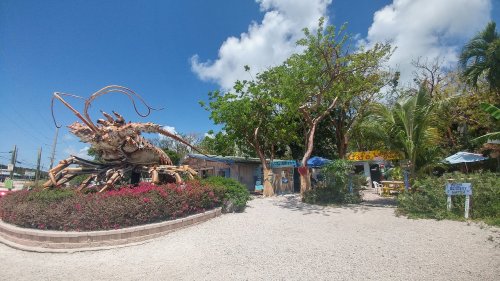 A large lobster sculpture stands outside a colorful café surrounded by greenery and blue skies.