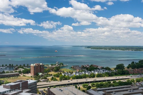 A scenic view of a lake with a blue sky, clouds, and a shoreline featuring buildings and greenery.