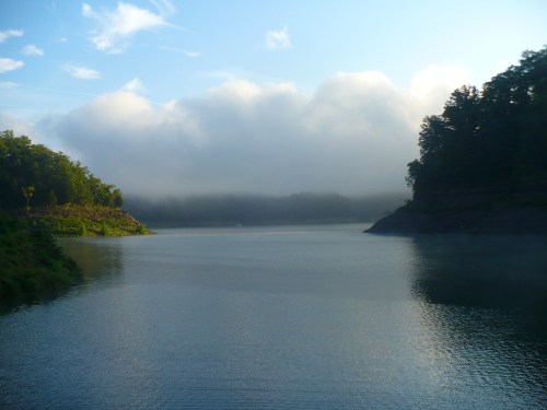 A serene lake surrounded by lush greenery and misty clouds, reflecting the calm water under a blue sky.
