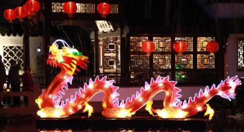 A brightly lit dragon lantern floats on water, surrounded by red lanterns and traditional architecture at night.