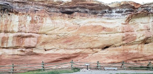Rocky cliff face with layered colors and textures, featuring a wooden fence and informational sign in the foreground.