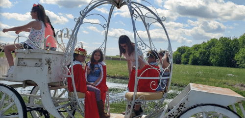Children dressed in costumes sit in a whimsical carriage, while another child rides on top, surrounded by greenery.