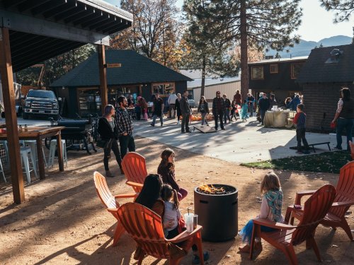 A group of people gather outdoors around a fire pit, with children sitting nearby and buildings in the background.