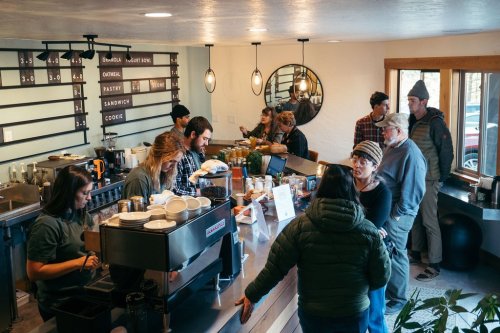 A busy café scene with baristas serving customers and people enjoying food and drinks at a communal space.