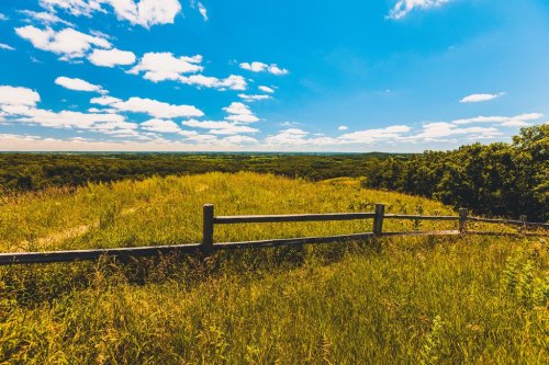 A scenic view of a grassy hill with a wooden fence, under a bright blue sky with fluffy clouds.