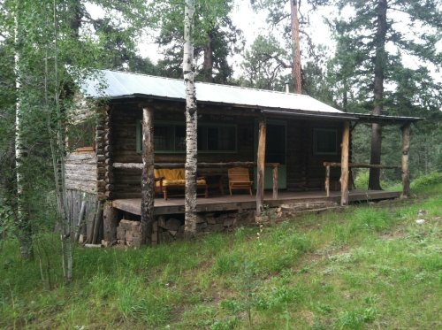 A rustic log cabin surrounded by trees, featuring a porch with two chairs and a metal roof.