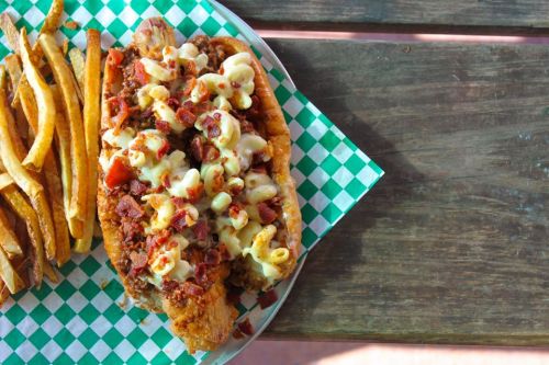 A loaded hot dog topped with macaroni and cheese, bacon bits, served with a side of golden fries on a checkered plate.