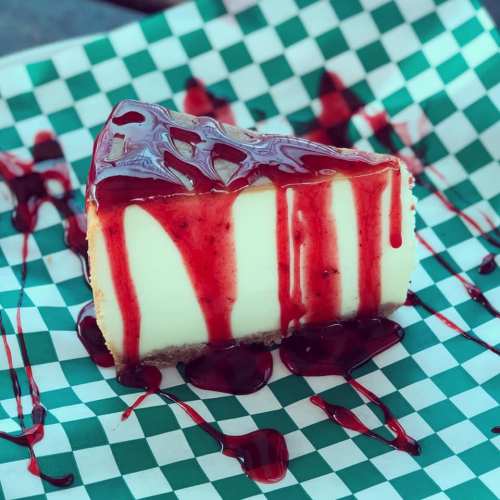 A slice of cheesecake drizzled with red berry sauce on a green and white checkered paper.