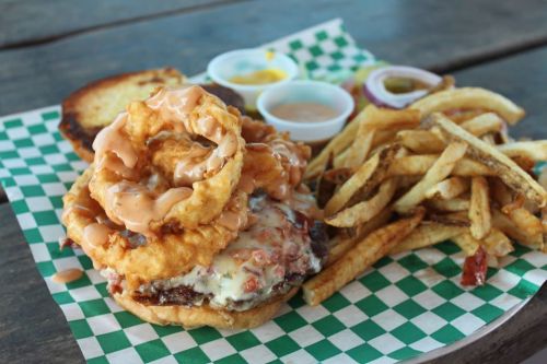 A hearty burger topped with onion rings and sauce, served with fries and dipping sauces on a checkered paper plate.