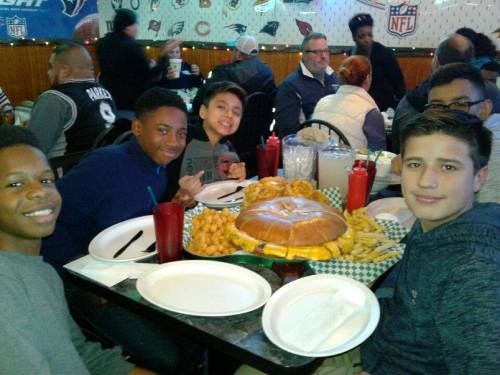 A group of boys smiles at the camera, surrounded by large plates of food in a lively restaurant setting.