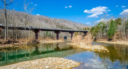 A bridge spans a clear river surrounded by trees and hills under a bright blue sky with scattered clouds.