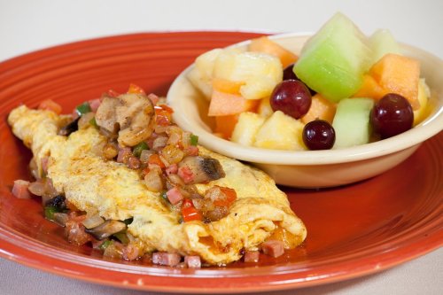 A colorful omelette topped with vegetables, served alongside a bowl of mixed fresh fruit.