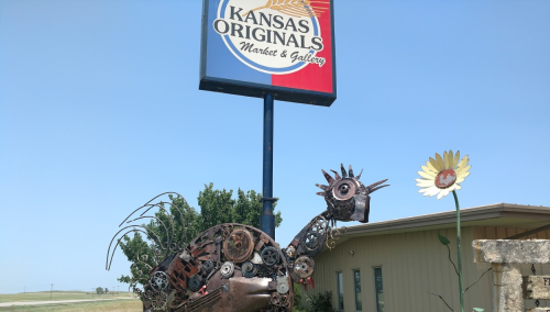 A metal sculpture of a bird stands beside a sign for Kansas Originals Market & Gallery, with a sunflower nearby.