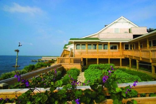 A wooden building by the water, surrounded by greenery and flowers, with a clear blue sky in the background.