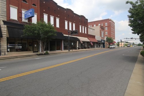 Empty street lined with brick buildings and storefronts, featuring a CPA sign and a few trees under a cloudy sky.