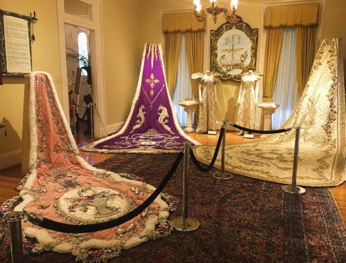 Three ornate, colorful ceremonial capes displayed in a museum setting, surrounded by a black rope barrier.