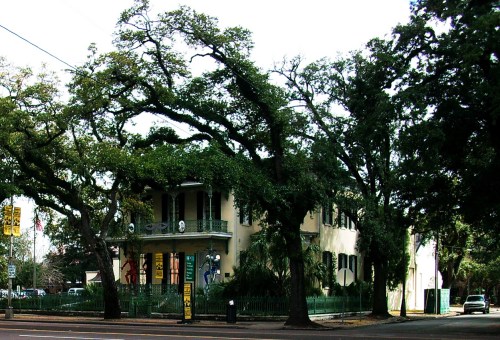Historic building surrounded by large trees, featuring colorful art on the exterior and a green fence.