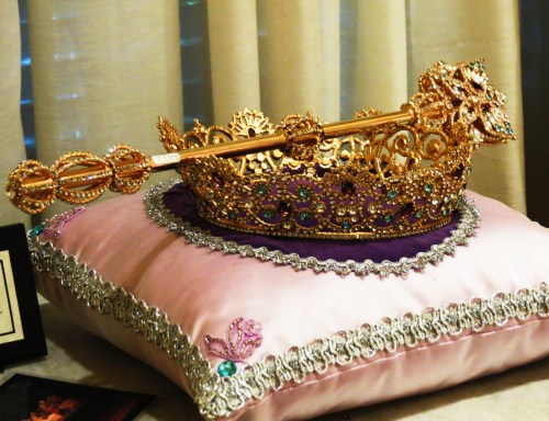 A golden crown adorned with jewels resting on a decorative pink pillow.