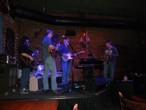 A live band performs on stage in a brick-walled venue, featuring guitars, keyboards, and a saxophone.