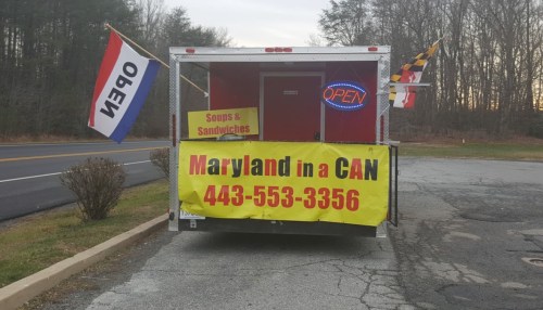 Food truck with a sign reading "Maryland in a CAN" and flags, offering soups and sandwiches, parked by the road.