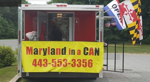 Food truck with a bright yellow banner reading "Maryland in a CAN" and flags, parked in a green area.