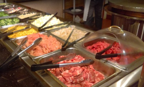 A buffet display with various meats and vegetables in trays, including chicken, beef, and assorted toppings.