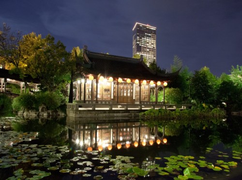 A traditional pavilion with lanterns reflects in a pond, surrounded by greenery, with a modern skyscraper in the background.