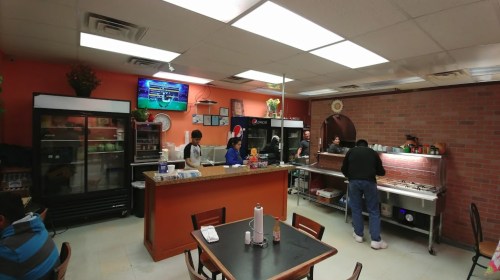 A cozy restaurant interior with staff preparing food, a TV on the wall, and tables for customers.