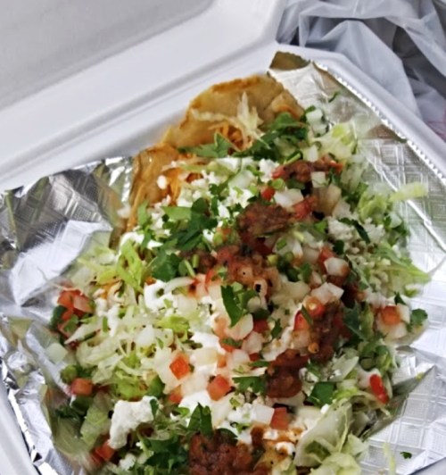 A takeout container with a taco topped with lettuce, tomatoes, onions, cilantro, and cheese on a bed of shredded lettuce.