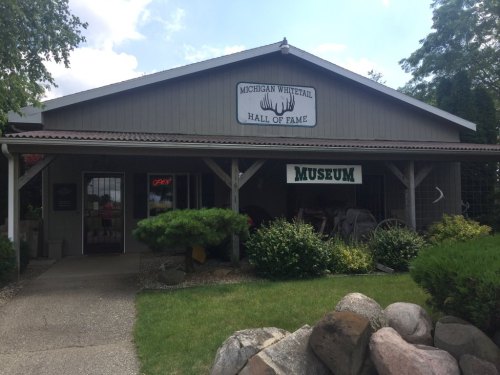 Exterior of the Michigan Whitetail Hall of Fame, featuring a sign and landscaped entrance with rocks and greenery.