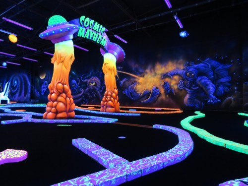 A vibrant indoor mini-golf course with glowing alien-themed decor and colorful obstacles under blacklight.