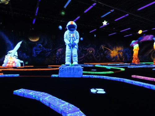 A glow-in-the-dark mini-golf course featuring an astronaut statue and colorful, illuminated pathways.