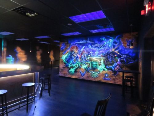 A vibrant mural featuring colorful characters and scenes on a brick wall in a dimly lit bar setting.