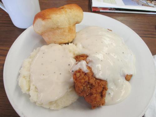 A plate with fried chicken covered in gravy, mashed potatoes, and a soft roll.