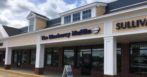 Exterior of a storefront featuring "The Blueberry Muffin" sign, with a light-colored building and blue sky.