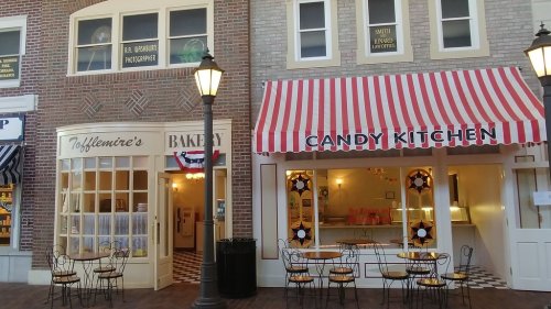 A quaint street scene featuring a bakery and a candy shop with colorful awnings and outdoor seating.