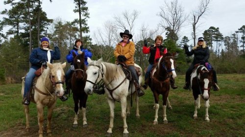 Five women on horseback, smiling and waving, surrounded by trees in a rural setting.